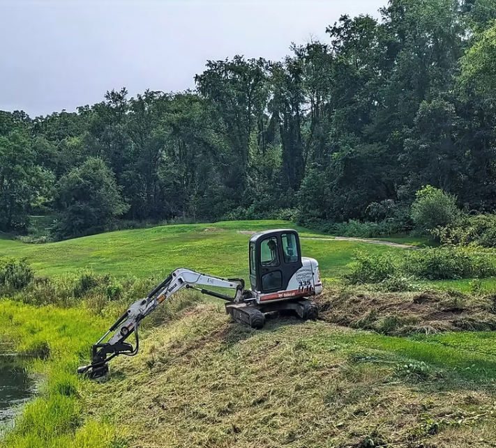Excavator performing vegetation maintenance beside a pond